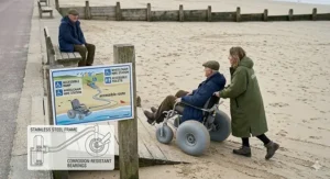 A person in a sand wheelchair with balloon tyres being pushed up a wooden accessible ramp at a UK beach with clear signage.
