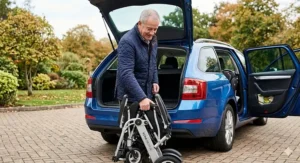 A senior man in a navy quilted jacket folding a modern, lightweight electric wheelchair on a British block-paving driveway next to an estate car with an open boot.