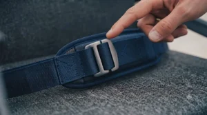 An adult finger points to the metallic tri-glide adjuster slide on a dark blue padded wheelchair safety belt, demonstrating how to adjust the strap length.