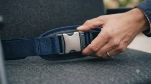 An adult hand with a simple band demonstrates the quick-release mechanism of the robust metallic buckle on a dark blue padded wheelchair lap strap.