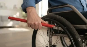 A user with limited dexterity operating a push-to-lock brake using a long red wheelchair wheel lock extension handle.