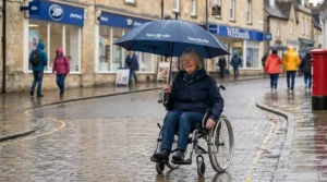 A person using a wheelchair fitted with a clamping umbrella, showing clear visibility and ventilation during light British drizzle.